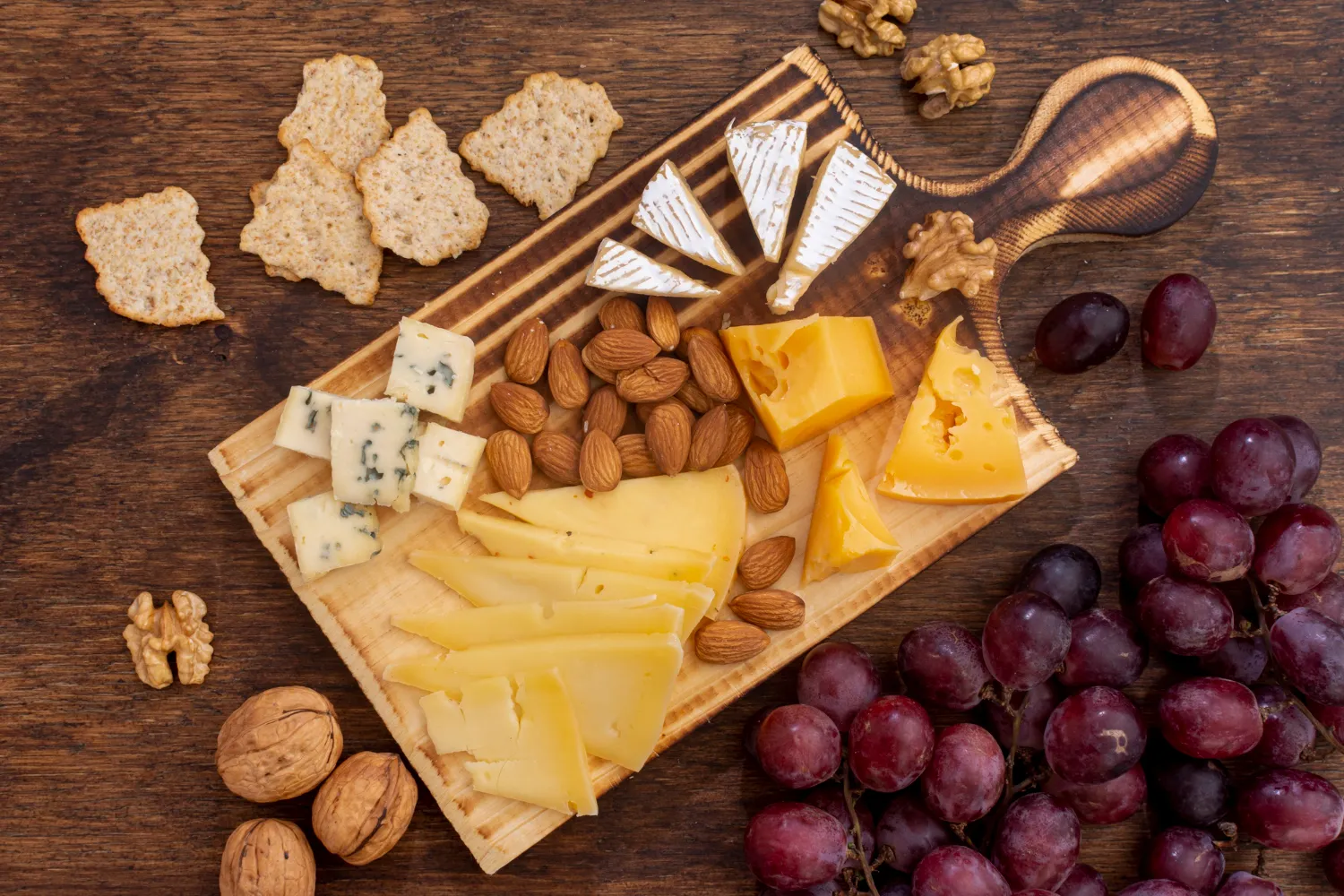 Tabla de quesos variados con almendras, nueces, uvas moradas y galletas sobre una superficie de madera. Ideal para una cena o evento social.