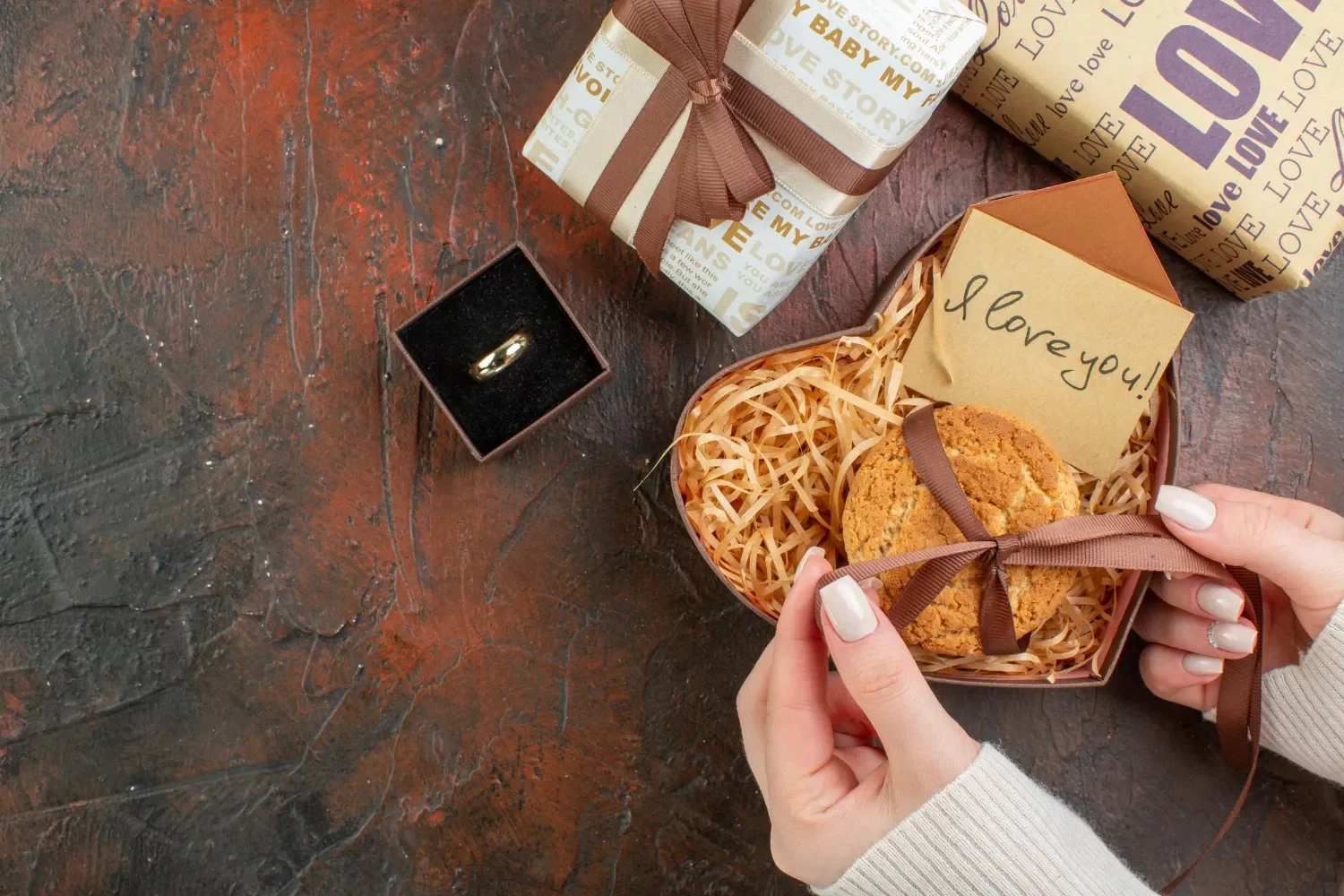 Manos sosteniendo una galleta decorada con un lazo, junto a cajas de regalo y un anillo en una caja negra, sobre una superficie oscura. Ideal para ocasiones especiales como aniversarios o propuestas.