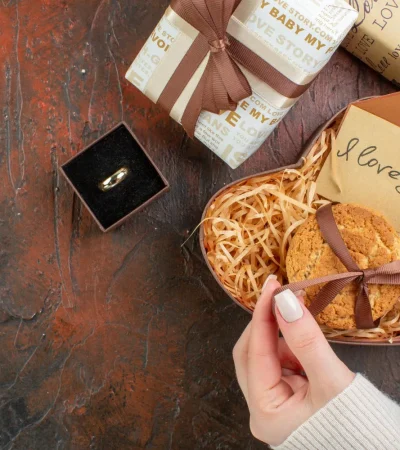 Manos sosteniendo una galleta decorada con un lazo, junto a cajas de regalo y un anillo en una caja negra, sobre una superficie oscura. Ideal para ocasiones especiales como aniversarios o propuestas.